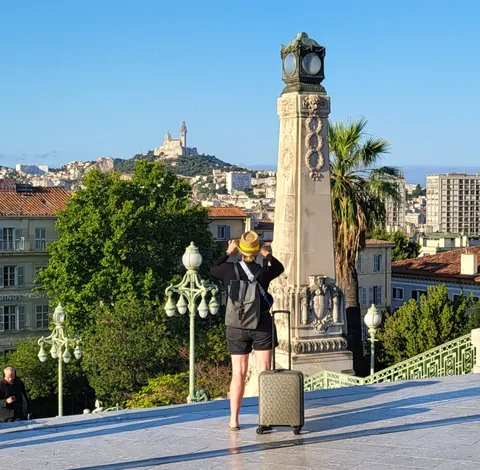 Une voyageuse avec sa valise sur une esplanade, Notre-Dame-de-la-Garde au loin