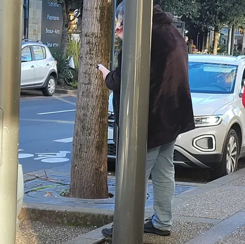 Un homme en manteau sombre à moitié caché derrière un tronc d’arbre et un poteau, au bord d’une rue