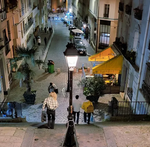 Une rue en pente bordée de cafés, le soir, un réverbère et des passants qui descendent les marches