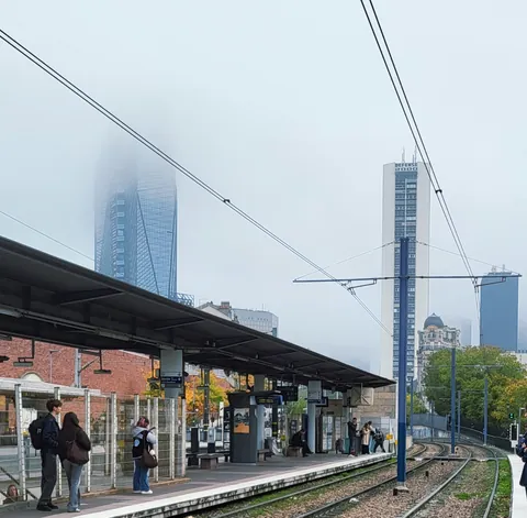Un quai de gare sous la brume, le haut des tours effacé par le brouillard