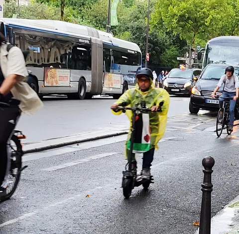 Un livreur en ciré jaune sur une trottinette, entre bus et vélos
