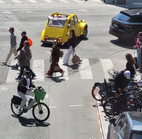 Une 2CV jaune au milieu d’un carrefour, des passants et des vélos