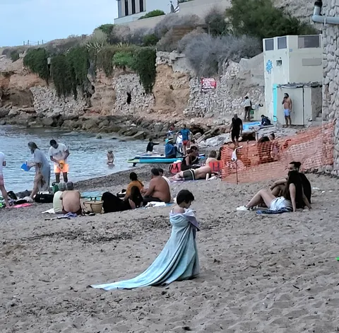 Une femme en longue robe bleue assise sur le sable d’une crique