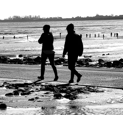 Deux silhouettes noires qui marchent sur une plage à marée basse, en noir et blanc