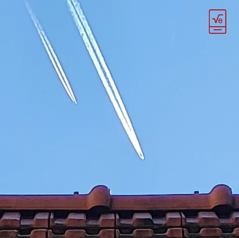 Deux traînées d’avion qui filent en diagonale dans le ciel bleu, au-dessus de toits de tuiles