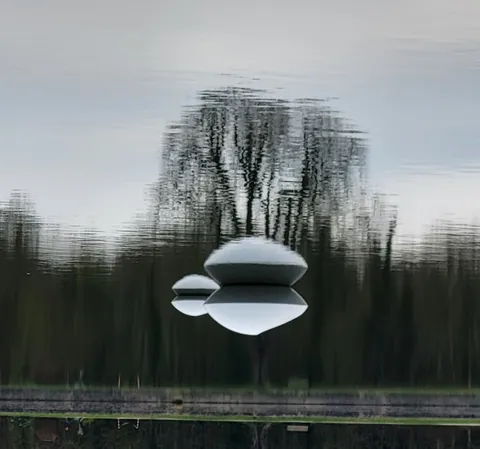 Une forme blanche de soucoupe volante qui flotte devant le reflet flou d’arbres dans l’eau