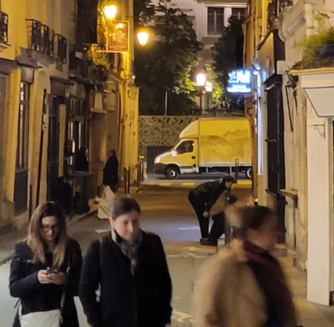Des passants dans une rue de nuit, une camionnette jaune au fond