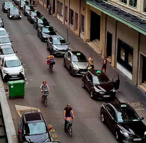 Une rue vue d’en haut, bordée de voitures garées, des cyclistes au milieu