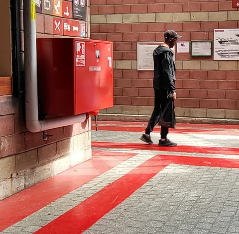 Un homme en noir qui marche sur un sol peint de bandes rouges, le long d’un mur de briques