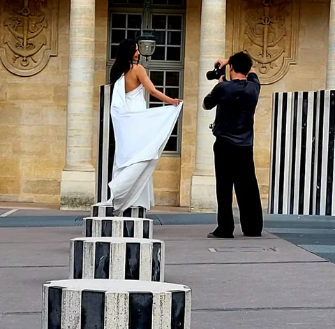 Une mariée en robe blanche posant sur une colonne de Buren face au photographe