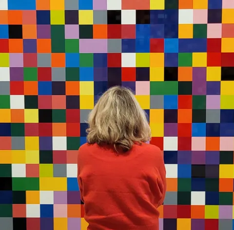 Une femme de dos devant un mur de carrés de couleur
