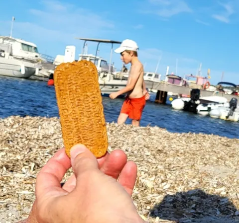 Une main qui tient un spéculoos devant une plage et un port, un enfant en maillot derrière
