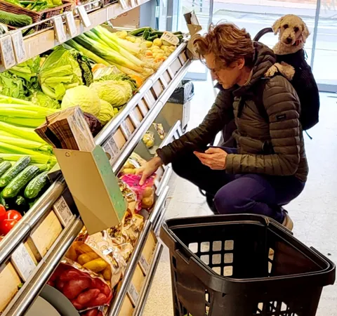 Une femme accroupie au rayon légumes, un caniche roux qui dépasse de son sac à dos