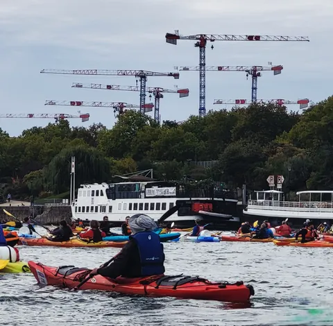 Des kayakistes sur un fleuve, une forêt de grues de chantier à l’arrière-plan