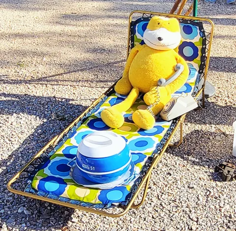 Une peluche jaune allongée sur un transat à fleurs, à côté d’un pot de crème